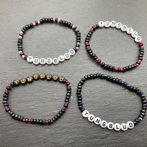 May include: Four beaded bracelets spelling "YUNGBLUD" in white letter beads. The bracelets are mainly black with pink, red, and blue accents. The bracelets are displayed on a dark grey surface.