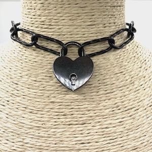 May include: A black choker necklace with a heart-shaped padlock pendant. The necklace has a chain-link design and is displayed on a woven, beige mannequin head. The padlock has a keyhole detail.