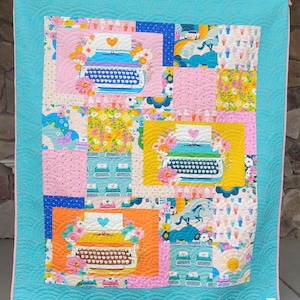 May include: A colorful quilt with a turquoise border and pink trim. The quilt features squares with floral and typewriter designs in pink, yellow, and orange. The quilt is machine-stitched with a wavy pattern.