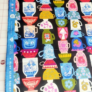 May include: Black fabric with a repeating pattern of stacked, colorful teacups and bowls. The teacups feature various designs, including Alice in Wonderland characters, flowers, and geometric shapes. The fabric's edge has text that reads "Ruby Star Society."