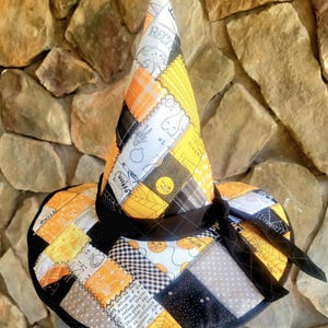 May include: A conical hat constructed from quilted fabric patches in black, white, orange, and yellow. The fabric features various patterns including spiderwebs, moons, and text. A black fabric band is tied around the base of the crown.