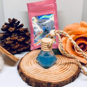 Puede incluir: Un ambientador de coche de cristal facetado con líquido azul, una tapa de madera y un cordón trenzado. El ambientador está sobre una rodaja de madera, con una bolsa rosa y una rosa en el fondo. La bolsa tiene texto.