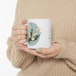 Puede incluir: Taza de cerámica blanca con una ilustración floral de flores de lirio de los valles en rosa y verde, sobre un óvalo azul claro. La taza tiene el texto "Lily of the Valley" en disposición vertical en el lado derecho.