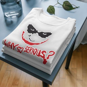 May include: A stack of white t-shirts featuring a graphic of a smiling face with black eye coverings and red lips. The text "WHY SO SERIOUS?" is printed in red below the face.