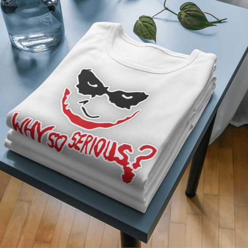 May include: A stack of white t-shirts featuring a graphic of a smiling face with black eye coverings and red lips. The text "WHY SO SERIOUS?" is printed in red below the face.