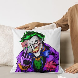 May include: White square pillow featuring a colorful illustration of the Joker character. The Joker is depicted with green hair, purple suit, and a sinister grin, holding a playing card. A pink flower is pinned to his lapel.