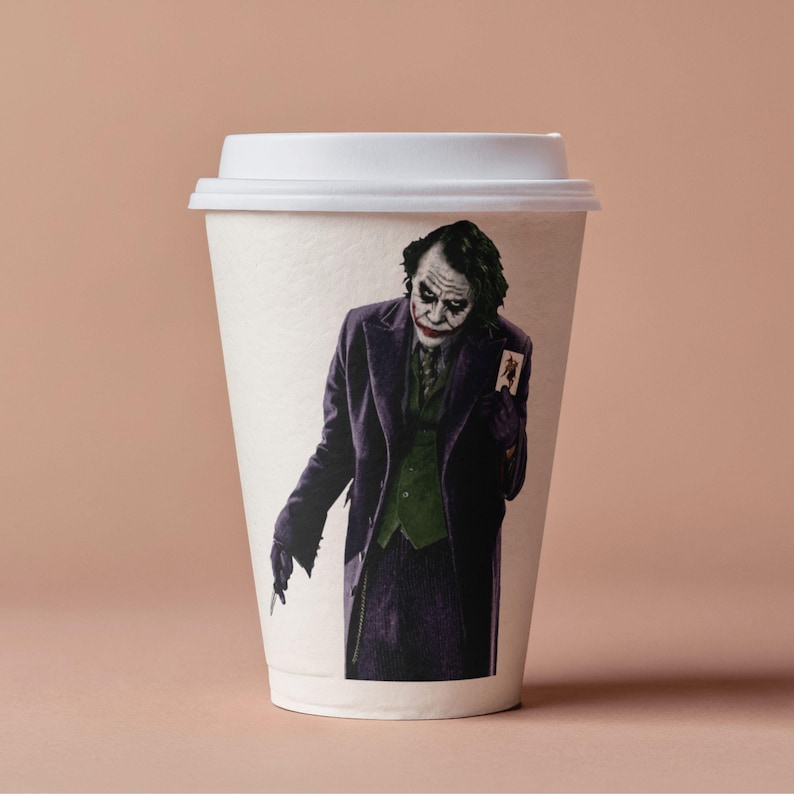 May include: A white paper coffee cup with a white lid. The cup features a full-color image of the Joker from the Batman movie, wearing a purple coat and green vest, holding a playing card. The background is a solid light brown.