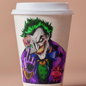 May include: A white paper coffee cup featuring a colorful illustration of the Joker from DC Comics. The Joker is depicted with green hair, white face paint, and a purple suit, holding a playing card. The design is a vibrant and detailed graphic.