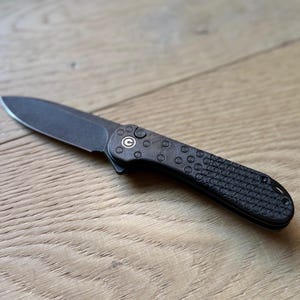 May include: A black folding knife with a textured black handle and a silver circle with a "C" inside. The knife is closed and lying on a light brown wooden surface.