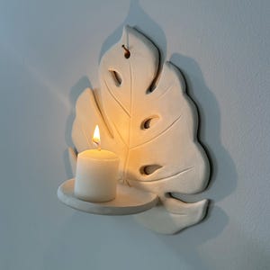Puede incluir: Un portavelas de cer&aacute;mica blanca montado en la pared con forma de hoja de Monstera. Una vela blanca encendida se encuentra sobre una peque&ntilde;a plataforma redonda. La llama de la vela brilla intensamente, proyectando sombras en la pared. El soporte tiene detalles recortados.