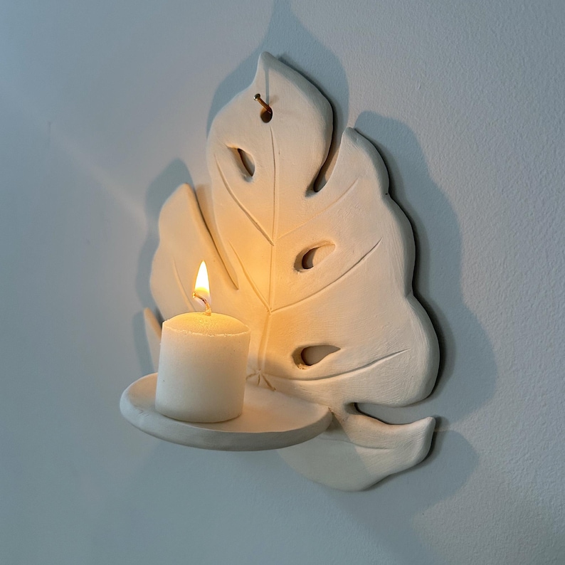 Puede incluir: Un portavelas de cer&aacute;mica blanca montado en la pared con forma de hoja de Monstera. Una vela blanca encendida se encuentra sobre una peque&ntilde;a plataforma redonda. La llama de la vela brilla intensamente, proyectando sombras en la pared. El soporte tiene detalles recortados.