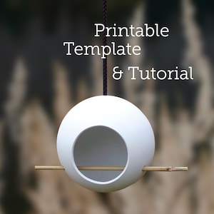 May include: White ceramic bird feeder shaped like a sphere with a round opening and a wooden perch. The feeder is hanging from a black cord and the text "Printable Template & Tutorial" is above the feeder.