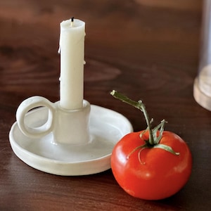 K&ouml;nnte beinhalten: Ein wei&szlig;er Keramik-Kerzenhalter mit Griff h&auml;lt eine brennende, cremefarbene Kerze. Eine reife rote Tomate mit gr&uuml;nem Stiel liegt daneben. Die Gegenst&auml;nde befinden sich auf einer dunklen Holzoberfl&auml;che.
