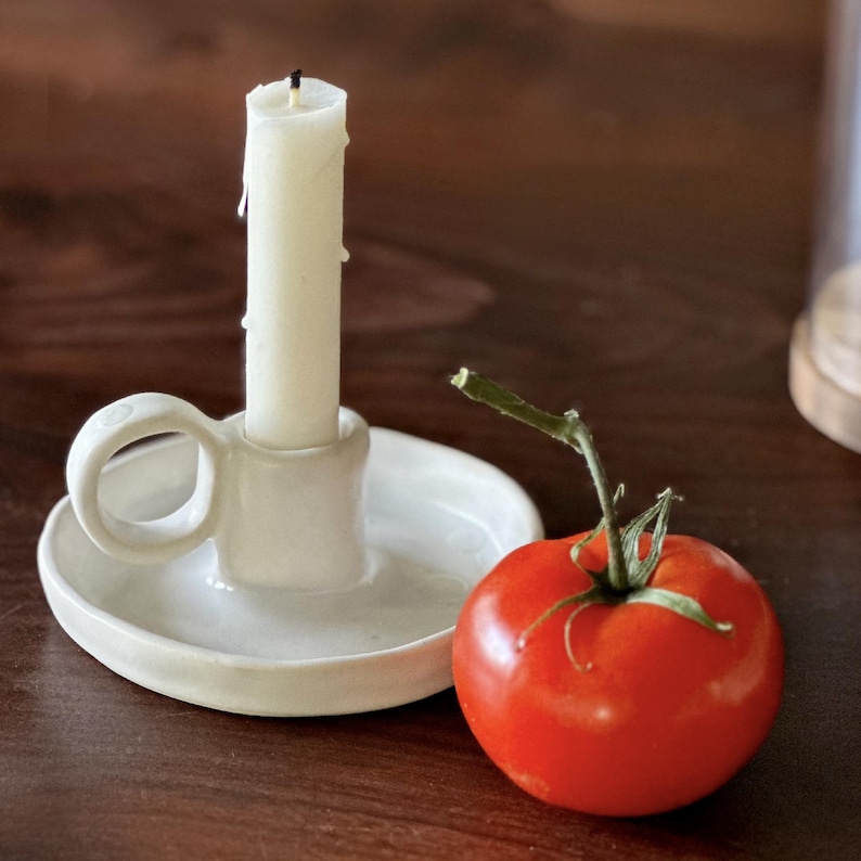 K&ouml;nnte beinhalten: Ein wei&szlig;er Keramik-Kerzenhalter mit Griff h&auml;lt eine brennende, cremefarbene Kerze. Eine reife rote Tomate mit gr&uuml;nem Stiel liegt daneben. Die Gegenst&auml;nde befinden sich auf einer dunklen Holzoberfl&auml;che.