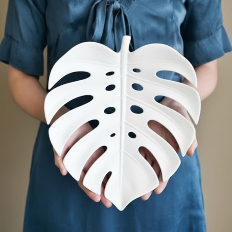May include: A white, leaf-shaped decorative plate with a monstera design. The plate features a central vein and several oval-shaped cutouts. The plate is held in front of a person wearing a blue top.