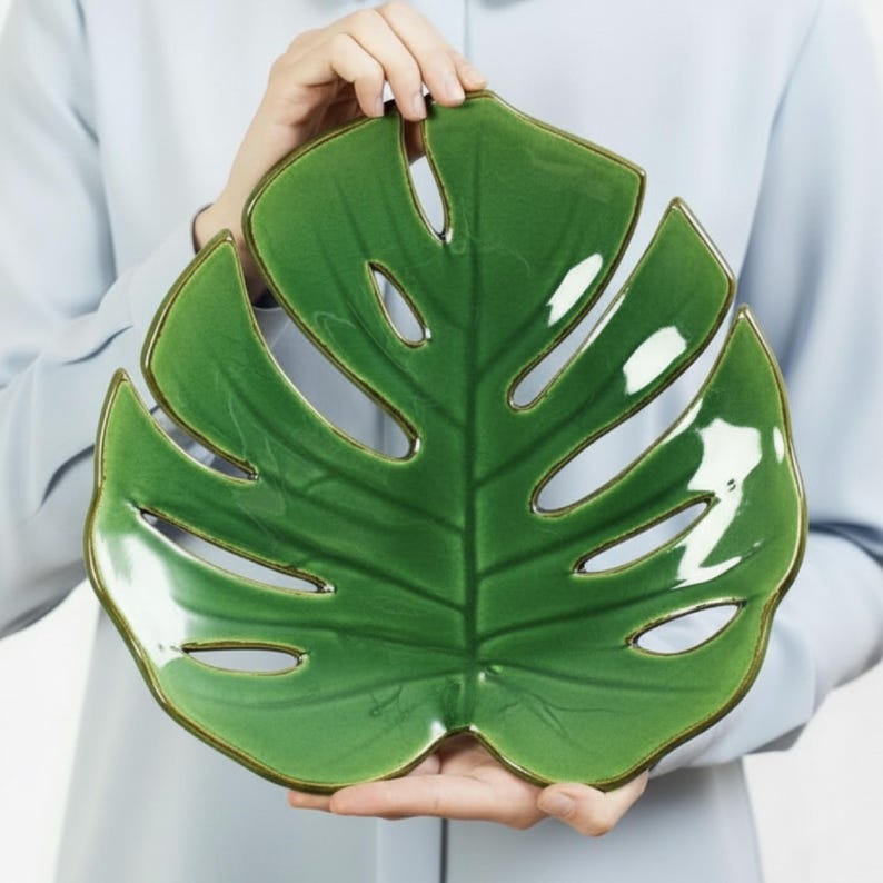 May include: A glossy green ceramic leaf-shaped serving dish with a brown rim. The dish has several oval cutouts and is held by a person wearing a light blue shirt. The dish is designed to resemble a Monstera leaf.