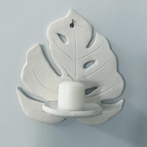 Puede incluir: Candelabro de pared de cer&aacute;mica blanca con forma de hoja de monstera. La hoja tiene detalles recortados y sostiene una vela de pilar blanca. El candelabro est&aacute; montado en una pared azul claro.