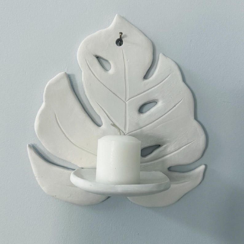 Puede incluir: Candelabro de pared de cer&aacute;mica blanca con forma de hoja de monstera. La hoja tiene detalles recortados y sostiene una vela de pilar blanca. El candelabro est&aacute; montado en una pared azul claro.