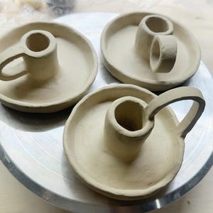K&ouml;nnte beinhalten: Drei unglasierte Kerzenhalter aus Keramik. Jeder hat eine kreisf&ouml;rmige Basis mit einem zentralen Zylinder f&uuml;r eine Kerze. Zwei haben einen Griff, und einer hat ein Doppelzylinder-Design. Die Keramik ist hellbeige.