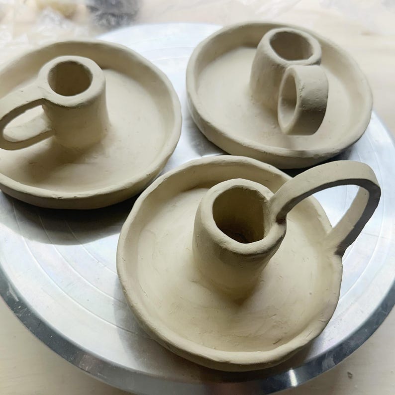 K&ouml;nnte beinhalten: Drei unglasierte Kerzenhalter aus Keramik. Jeder hat eine kreisf&ouml;rmige Basis mit einem zentralen Zylinder f&uuml;r eine Kerze. Zwei haben einen Griff, und einer hat ein Doppelzylinder-Design. Die Keramik ist hellbeige.