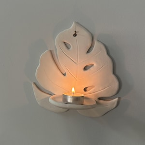 Puede incluir: Un aplique de pared blanco con forma de hoja con un peque&ntilde;o portavelas redondo. Una vela de t&eacute; encendida est&aacute; en el soporte, proyectando un brillo c&aacute;lido. El aplique est&aacute; montado en una pared gris claro, creando una est&eacute;tica minimalista.
