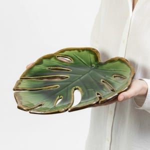 May include: A green ceramic leaf-shaped serving dish with a glossy finish and brown trim. The dish has several cut-out sections and is held by a person wearing a white shirt. The design mimics a Monstera plant leaf.