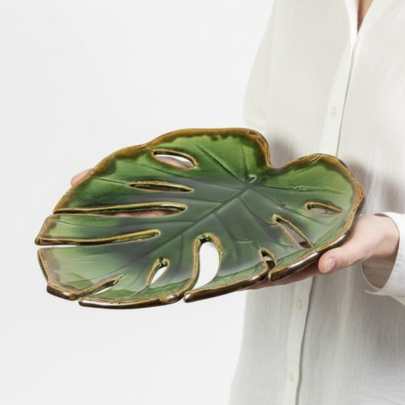 May include: A green ceramic leaf-shaped serving dish with a glossy finish and brown trim. The dish has several cut-out sections and is held by a person wearing a white shirt. The design mimics a Monstera plant leaf.