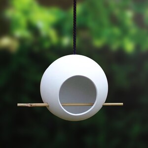 Puede incluir: Comedero para p&aacute;jaros de cer&aacute;mica blanca con forma de huevo, con una abertura redonda y una percha de madera. El comedero cuelga de un cord&oacute;n negro.