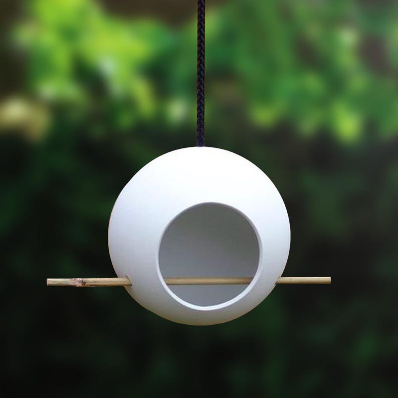 Puede incluir: Comedero para p&aacute;jaros de cer&aacute;mica blanca con forma de huevo, con una abertura redonda y una percha de madera. El comedero cuelga de un cord&oacute;n negro.