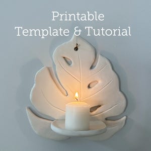 May include: A white, leaf-shaped wall sconce with a lit white candle. The sconce has a monstera leaf design and includes a small, round candle holder. The text "Printable Template & Tutorial" is at the top.