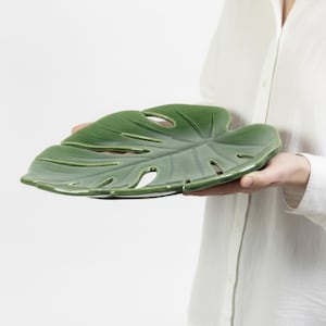 May include: A green ceramic plate in the shape of a Monstera leaf. The plate has a glossy finish and features cut-out holes, mimicking the natural leaf's appearance. The plate is held by a person wearing a white shirt.