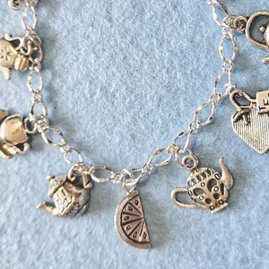 May include: Silver-toned charm bracelet featuring tea-themed charms. Charms include teacups, teapots, a lemon slice, and a tag with the word "TEA". The bracelet has a lobster clasp closure and is on a light blue background.