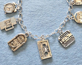 May include: Silver-toned charm bracelet featuring various vintage-themed charms. Charms include a clock, radio, television, typewriter, cassette tape, and a jukebox with the word "Juke Box". The bracelet is on a light blue background.