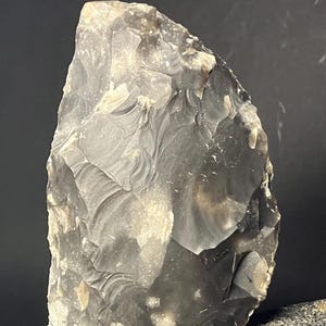 Puede incluir: Una herramienta de piedra grande y de forma tosca, con una paleta de colores gris oscuro y gris claro. La piedra tiene una superficie texturizada con escamas y astillas visibles, lo que sugiere que fue trabajada a mano. El objeto es probablemente un artefacto prehistórico.