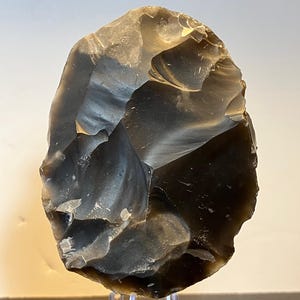 May include: A large, dark grey flint hand axe, oval in shape, with a rough, textured surface. The stone tool is displayed on a clear acrylic stand, showcasing its prehistoric craftsmanship. The axe exhibits natural variations in colour and texture.
