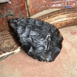 Puede incluir: Una herramienta de piedra de obsidiana negra con un borde afilado y escamado y una superficie lisa y brillante. La piedra presenta un patrón de fractura concoidea distintivo en su cara, mostrando un aspecto oscuro y reflectante con inclusiones grises.