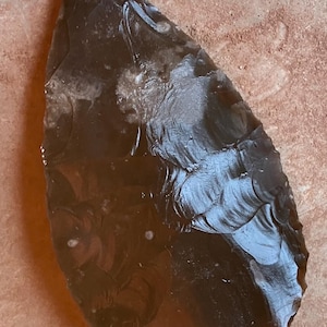 May include: A dark brown, teardrop-shaped stone tool with a polished surface. The tool has a sharp edge and appears to be hand-crafted. The stone has a translucent quality, with visible internal patterns. It rests on a rough, light brown surface.