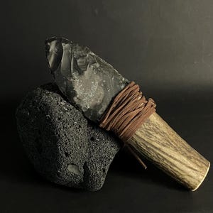 May include: A primitive flint knife with a wooden handle wrapped in brown leather cord rests on a dark grey, porous stone. The blade is a dark grey colour, and the handle has a natural wood grain.