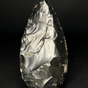 Peut inclure: Une pointe de flèche en obsidienne de couleur gris foncé, en forme de goutte d'eau. La pierre présente une surface polie avec des stries visibles et une pointe acérée. Outil préhistorique, sur un support transparent.