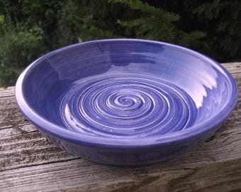 May include: A round, glazed ceramic bowl in a vibrant periwinkle blue color. The interior surface of the bowl features a distinct, raised spiral pattern that radiates from the center outward toward the smooth, curved edges of the vessel.