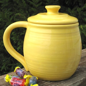 Pode incluir: Uma caneca de cerâmica amarela brilhante com tampa e alça a condizer. A caneca tem formato arredondado com ranhuras horizontais. Vários doces embalados em vermelho, roxo e azul estão ao lado da caneca. A caneca está sobre uma superfície de madeira.