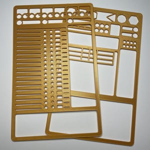 May include: Three gold-colored plastic stencils with various geometric shapes and measurement markings. The stencils are designed with multiple cut-out shapes, including circles, rectangles, and lines, for crafting or design projects. The stencils are stacked on a white surface.