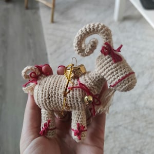 Peut inclure: Une décoration de chèvre en crochet avec une cloche dorée et un charme de boîte dorée. La chèvre est beige avec des accents rouges et une corne recourbée.