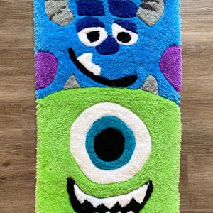 Può includere: Un tappeto rettangolare con due personaggi di mostri dei cartoni animati. La metà superiore è blu con una macchia viola e corna grigie, e la metà inferiore è verde con un grande occhio bianco e un sorriso nero. Il tappeto è realizzato in un materiale morbido e strutturato.