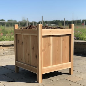 May include: A rectangular wooden planter box made of light-colored wood. The planter has a square shape with vertical wooden planks and a top filled with mulch. The box is outdoors on a stone patio.