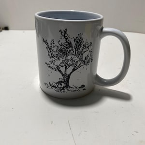 May include: White ceramic mug with a black and white illustration of a tree with a person sitting under it.