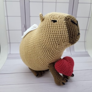 May include: A crocheted capybara plushie in tan with a brown face and black eyes. It has a small white wing on its back and is holding a red heart. The plushie is handmade.