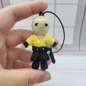 Pode incluir: Pequena boneca de crochê com camisa amarela, calças pretas e uma espada pequena. A boneca tem um tom de pele claro e um cordão preto para pendurar. Os braços da boneca têm listras marrons. A boneca tem cerca de 7.6 cm de altura.