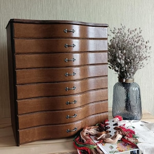 May include: A wooden chest with eight drawers and curved edges. The drawers have black metal handles. The chest is sitting on a wooden surface.