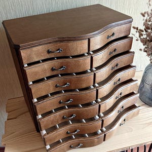 May include: A wooden jewelry chest with a dark brown finish. The chest has a curved top and features multiple drawers with decorative handles. The drawers are partially open, revealing the interior. The chest is placed on a wooden surface.